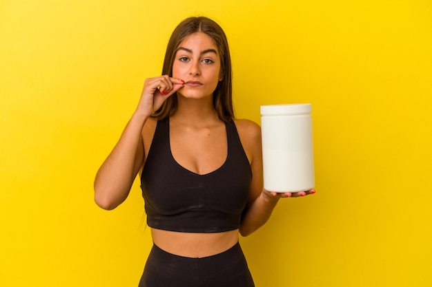 Joven mujer caucásica sosteniendo una botella de proteína aislada sobre fondo amarillo con los dedos en los labios guardando un secreto.