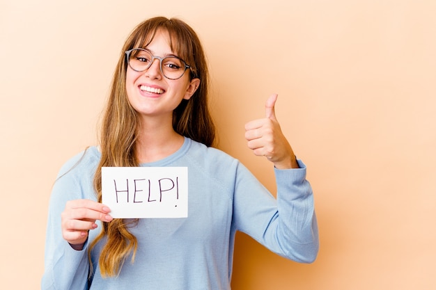 Joven mujer caucásica sosteniendo un cartel de ayuda aislado sonriendo y levantando el pulgar hacia arriba