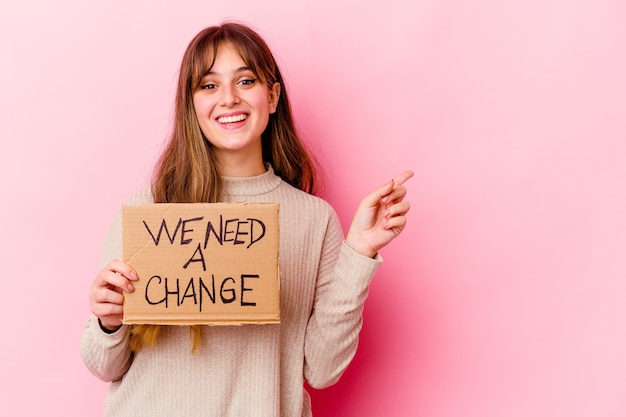 Joven mujer caucásica sosteniendo un cartel de Necesitamos un cambio aislado sonriendo y apuntando a un lado, mostrando algo en el espacio en blanco.