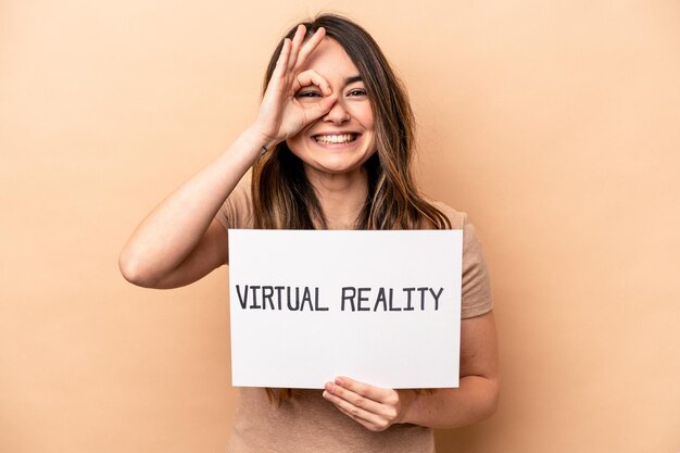 Joven mujer caucásica sosteniendo un cartel de realidad virtual aislado en un fondo beige emocionada manteniendo el gesto correcto en el ojo