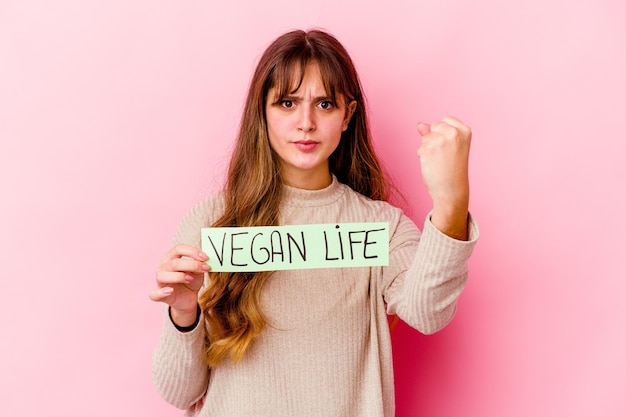 Foto joven mujer caucásica sosteniendo un cartel de vida vegana aislado mostrando el puño a la cámara, expresión facial agresiva.