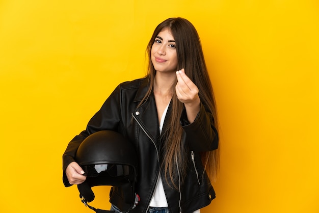 Joven mujer caucásica sosteniendo un casco de motocicleta aislado en la pared amarilla haciendo gesto de dinero