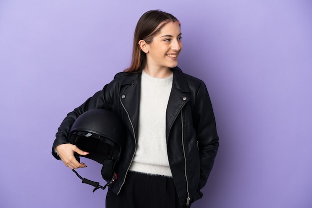 Joven mujer caucásica sosteniendo un casco de motocicleta aislado en la pared púrpura mirando hacia el lado