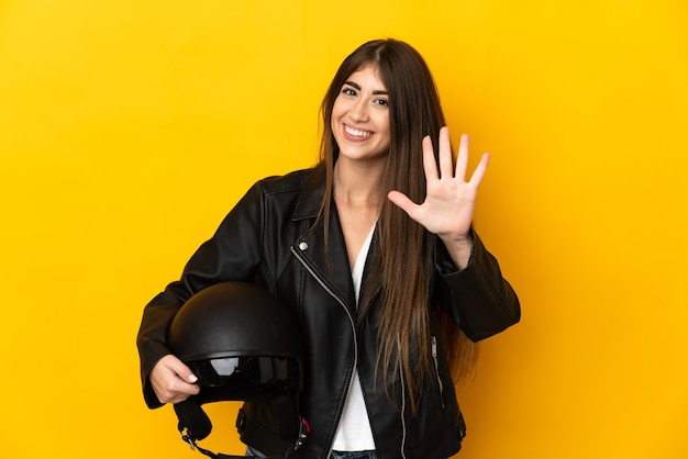 Joven mujer caucásica sosteniendo un casco de motocicleta aislado sobre fondo amarillo contando cinco con los dedos
