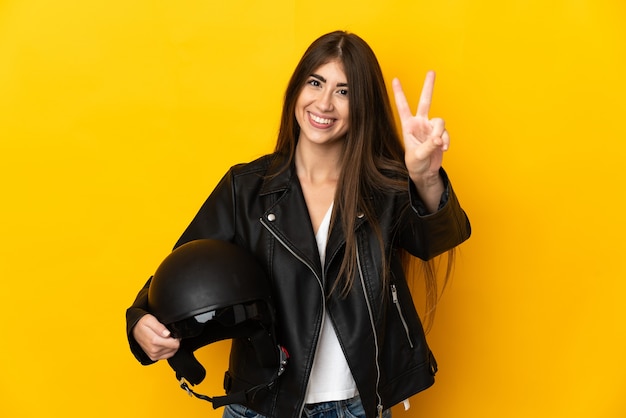 Joven mujer caucásica sosteniendo un casco de motocicleta aislado sobre fondo amarillo sonriendo y mostrando el signo de la victoria