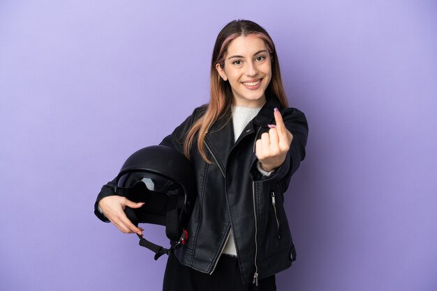 Joven mujer caucásica sosteniendo un casco de motocicleta aislado sobre fondo púrpura haciendo el gesto que viene