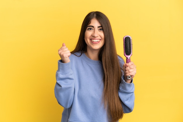 Joven mujer caucásica sosteniendo cepillo para el cabello aislado sobre fondo azul celebrando una victoria en la posición ganadora