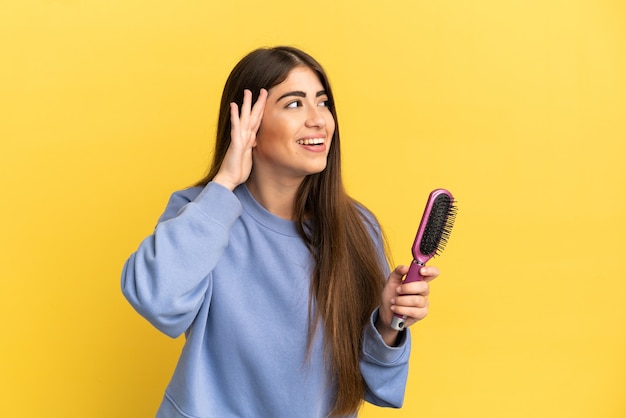 Joven mujer caucásica sosteniendo el cepillo para el cabello aislado sobre fondo azul escuchando algo poniendo la mano en la oreja