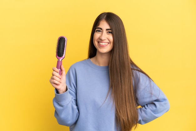 Joven mujer caucásica sosteniendo el cepillo para el cabello aislado sobre fondo azul posando con los brazos en la cadera y sonriendo