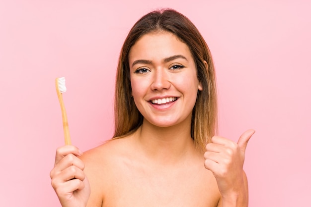 Joven mujer caucásica sosteniendo un cepillo de dientes aislado sonriendo y levantando el pulgar hacia arriba