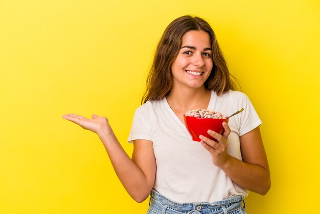 Joven mujer caucásica sosteniendo cereales aislado sobre fondo amarillo mostrando un espacio de copia en una palma y sosteniendo otra mano en la cintura.