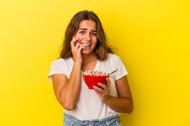 Joven mujer caucásica sosteniendo cereales aislados sobre fondo amarillo mordiéndose las uñas, nerviosa y muy ansiosa.