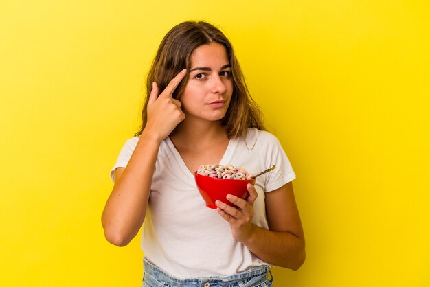 Joven mujer caucásica sosteniendo cereales aislados sobre fondo amarillo señalando el templo con el dedo, pensando, centrado en una tarea.