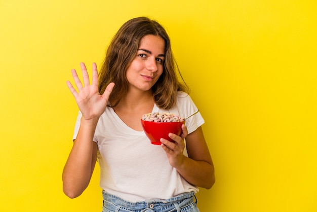 Joven mujer caucásica sosteniendo cereales aislados sobre fondo amarillo sonriendo alegre mostrando el número cinco con los dedos.