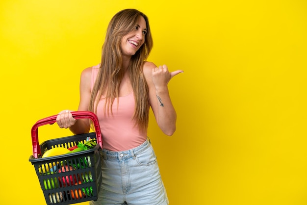Joven mujer caucásica sosteniendo una cesta de la compra llena de comida aislada en un fondo amarillo apuntando hacia un lado para presentar un producto