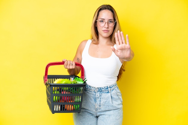 Joven mujer caucásica sosteniendo una cesta de la compra llena de comida aislada en un fondo amarillo haciendo un gesto de parada