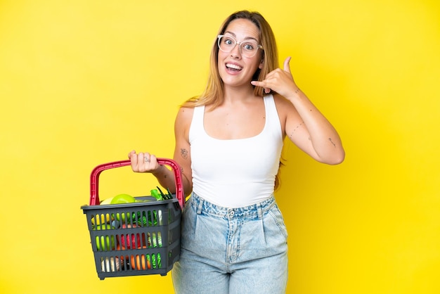 Joven mujer caucásica sosteniendo una cesta de la compra llena de comida aislada en un fondo amarillo haciendo un gesto telefónico Llámame de nuevo