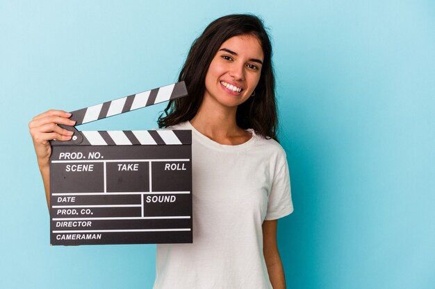 Joven mujer caucásica sosteniendo una claqueta aislada sobre fondo azul feliz, sonriente y alegre.