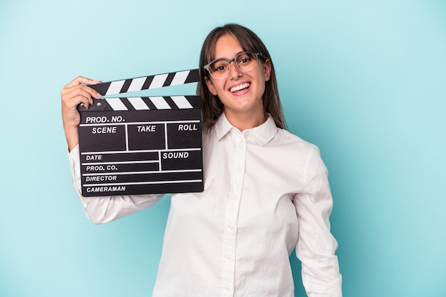 Joven mujer caucásica sosteniendo claqueta aislado sobre fondo azul feliz, sonriente y alegre.