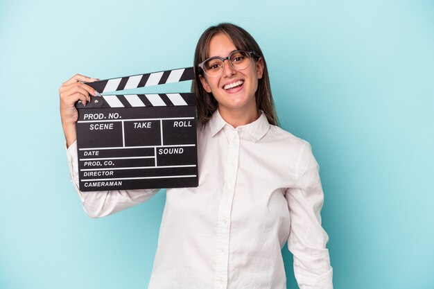 Joven mujer caucásica sosteniendo claqueta aislado sobre fondo azul feliz, sonriente y alegre.