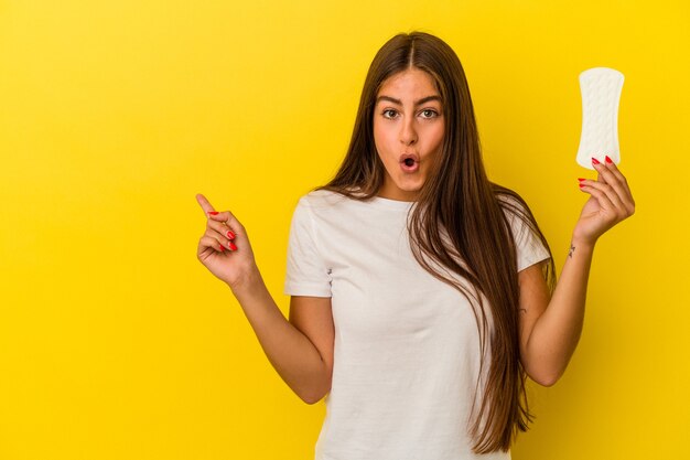 Joven mujer caucásica sosteniendo una compresa aislada sobre fondo amarillo apuntando hacia el lado