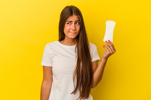 Joven mujer caucásica sosteniendo una compresa aislada sobre fondo amarillo confundida, se siente dudosa e insegura.