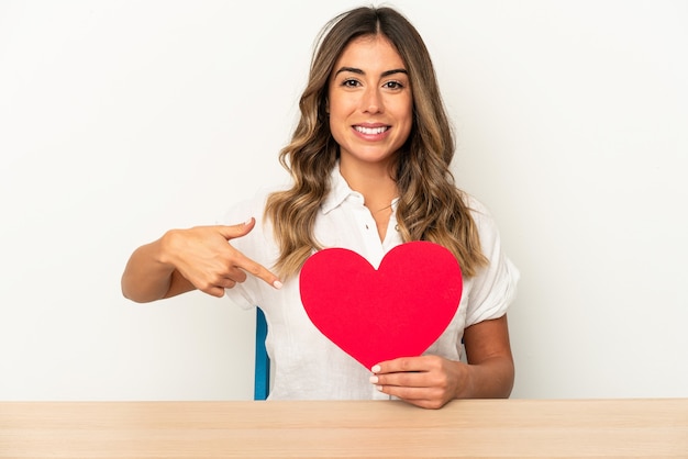 Joven mujer caucásica sosteniendo un corazón de San Valentín persona aislada apuntando con la mano a un espacio de copia de camisa, orgulloso y seguro