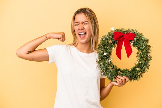 Joven mujer caucásica sosteniendo una corona de Navidad aislada sobre fondo amarillo levantando el puño después de una victoria, concepto ganador.