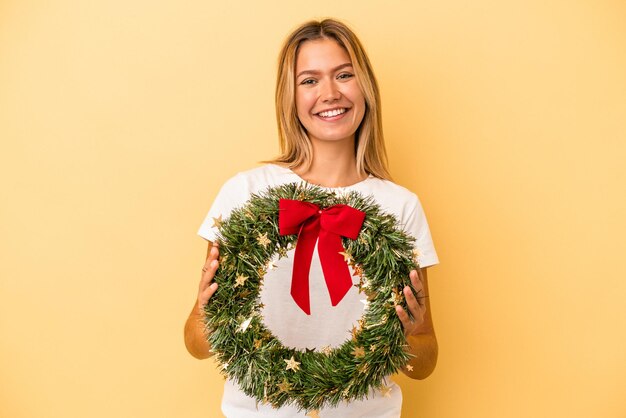 Joven mujer caucásica sosteniendo una corona de Navidad aislada sobre fondo amarillo