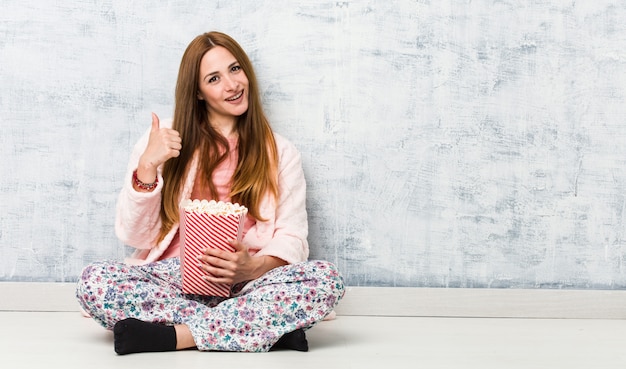 Joven mujer caucásica sosteniendo un cubo de palomitas de maíz sonriendo y levantando el pulgar