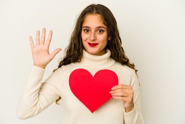 Joven mujer caucásica sosteniendo una forma de día de San Valentín de corazón aislada sonriendo alegre mostrando el número cinco con los dedos.