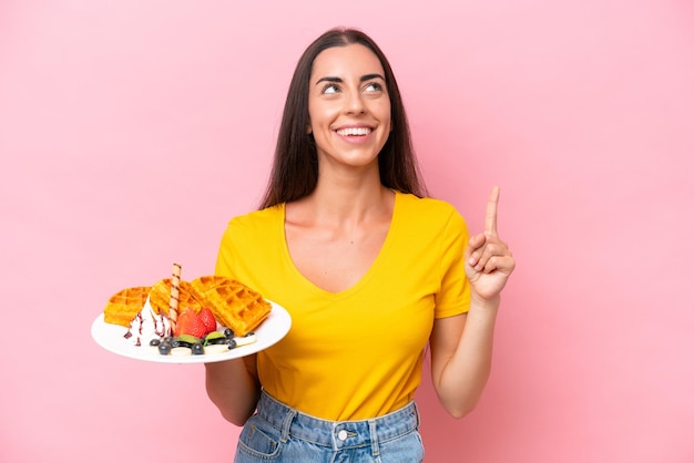 Joven mujer caucásica sosteniendo gofres aislados en un fondo rosado señalando una gran idea