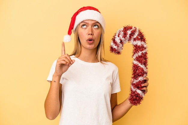 Joven mujer caucásica sosteniendo gran palo de Navidad aislado sobre fondo amarillo apuntando al revés con la boca abierta.