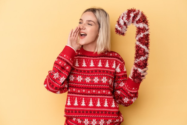 Joven mujer caucásica sosteniendo gran palo de Navidad aislado sobre fondo amarillo gritando y sosteniendo la palma cerca de la boca abierta.
