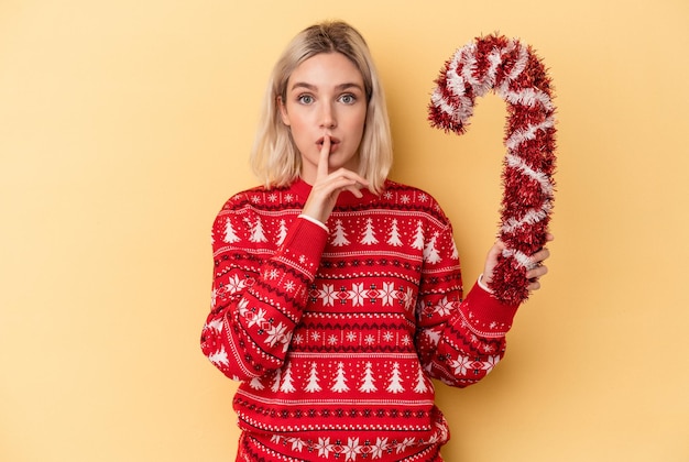 Joven mujer caucásica sosteniendo gran palo de Navidad aislado sobre fondo amarillo manteniendo un secreto o pidiendo silencio.