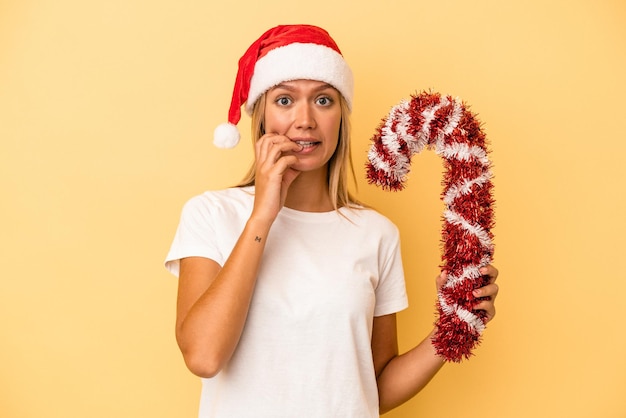 Joven mujer caucásica sosteniendo gran palo de Navidad aislado sobre fondo amarillo mordiéndose las uñas, nervioso y muy ansioso.