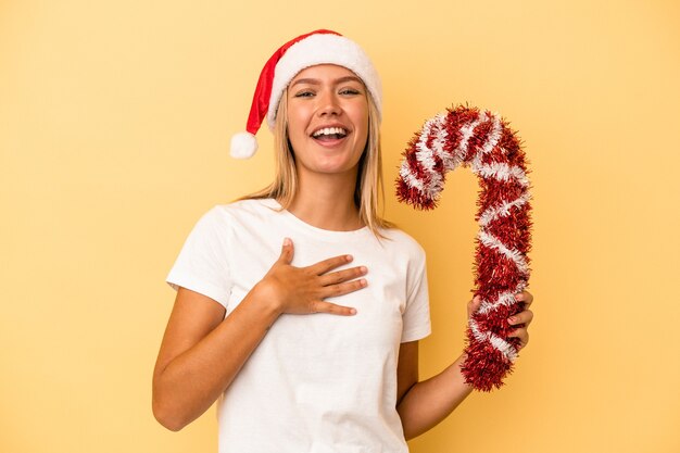 Joven mujer caucásica sosteniendo un gran palo de Navidad aislado sobre fondo amarillo se ríe a carcajadas manteniendo la mano en el pecho.