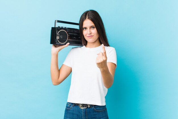 Joven mujer caucásica sosteniendo un guetto blaster apuntando con el dedo hacia usted como invitando a acercarse.