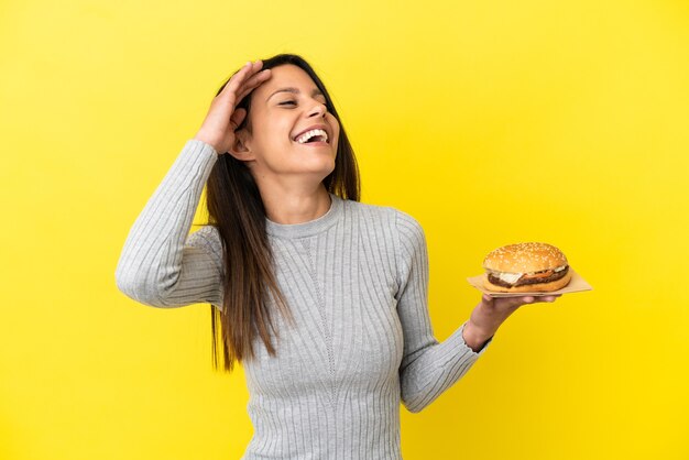 Joven mujer caucásica sosteniendo una hamburguesa aislada sobre fondo amarillo sonriendo mucho