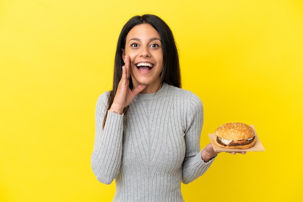 Joven mujer caucásica sosteniendo una hamburguesa aislada sobre fondo amarillo con sorpresa y expresión facial conmocionada