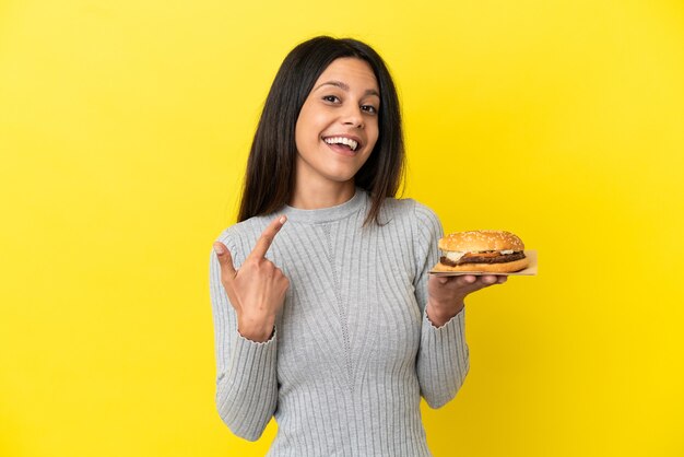 Joven mujer caucásica sosteniendo una hamburguesa aislado sobre fondo amarillo dando un pulgar hacia arriba gesto