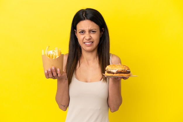 Joven mujer caucásica sosteniendo hamburguesas y patatas fritas aislado sobre fondo amarillo