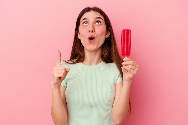 Joven mujer caucásica sosteniendo un helado aislado sobre fondo azul apuntando al revés con la boca abierta.