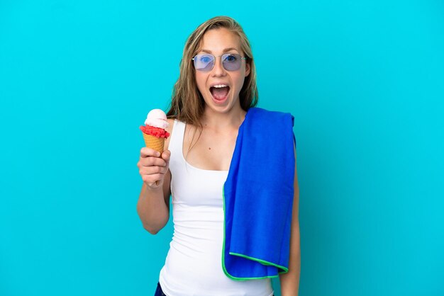 Joven mujer caucásica sosteniendo helado y usando una toalla de playa aislada de fondo azul con expresión facial sorprendida y sorprendida