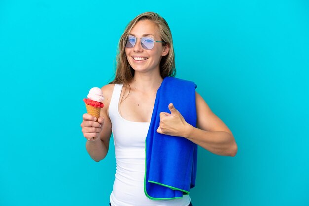 Joven mujer caucásica sosteniendo helado y usando una toalla de playa aislada de fondo azul con los pulgares hacia arriba porque algo bueno ha sucedido