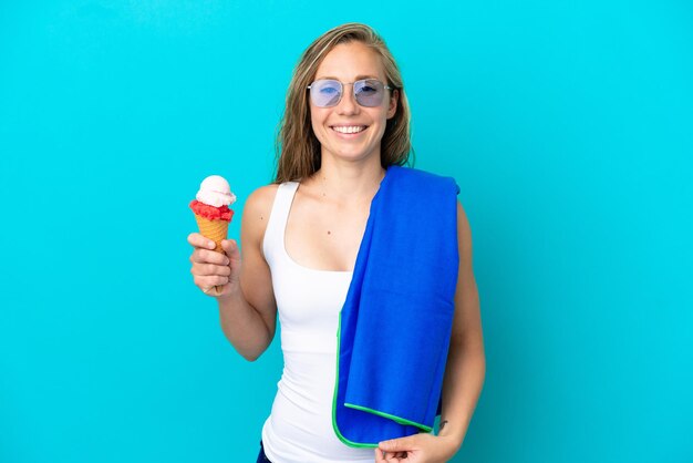 Joven mujer caucásica sosteniendo un helado y vistiendo una toalla de playa aislada sobre fondo azul sonriendo mucho