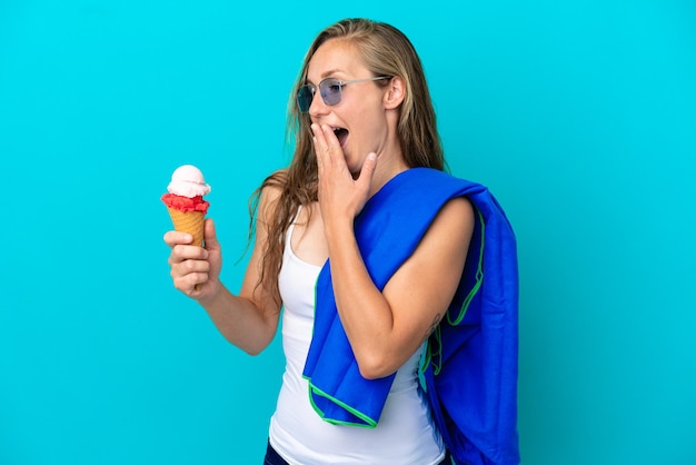 Joven mujer caucásica sosteniendo un helado y vistiendo una toalla de playa aislada sobre fondo azul con sorpresa y expresión facial conmocionada