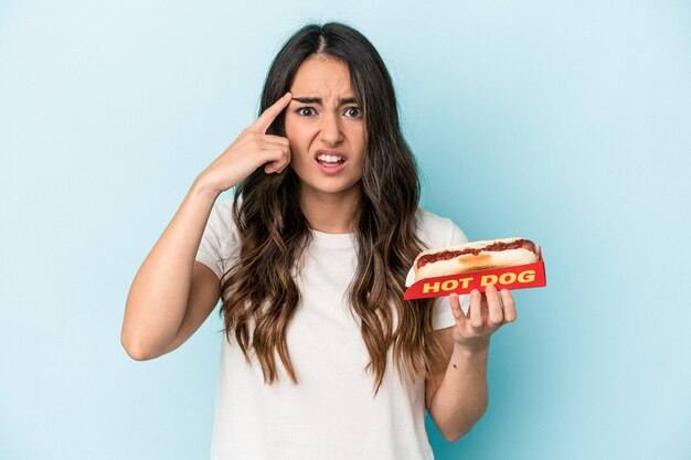 Joven mujer caucásica sosteniendo un hot dog aislado sobre fondo azul mostrando un gesto de decepción con el dedo índice.