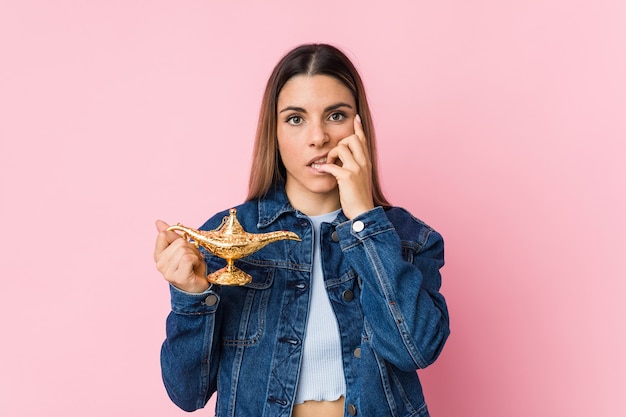 Joven mujer caucásica sosteniendo una lámpara mágica mordiéndose las uñas, nerviosa y muy ansiosa.