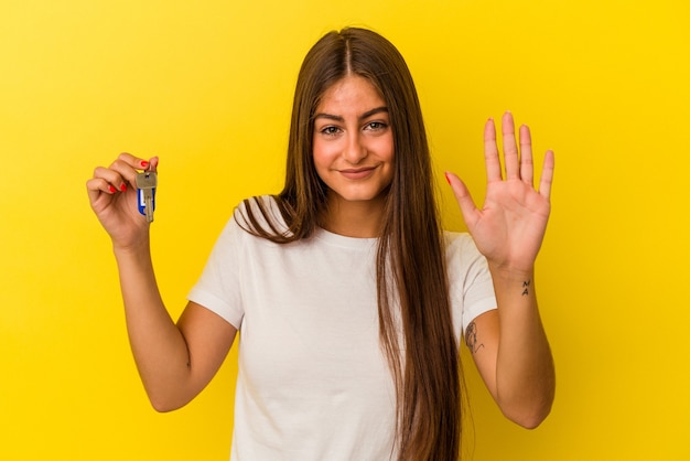 Joven mujer caucásica sosteniendo una llave de casa aislada en la pared amarilla sonriendo alegre mostrando el número cinco con los dedos.